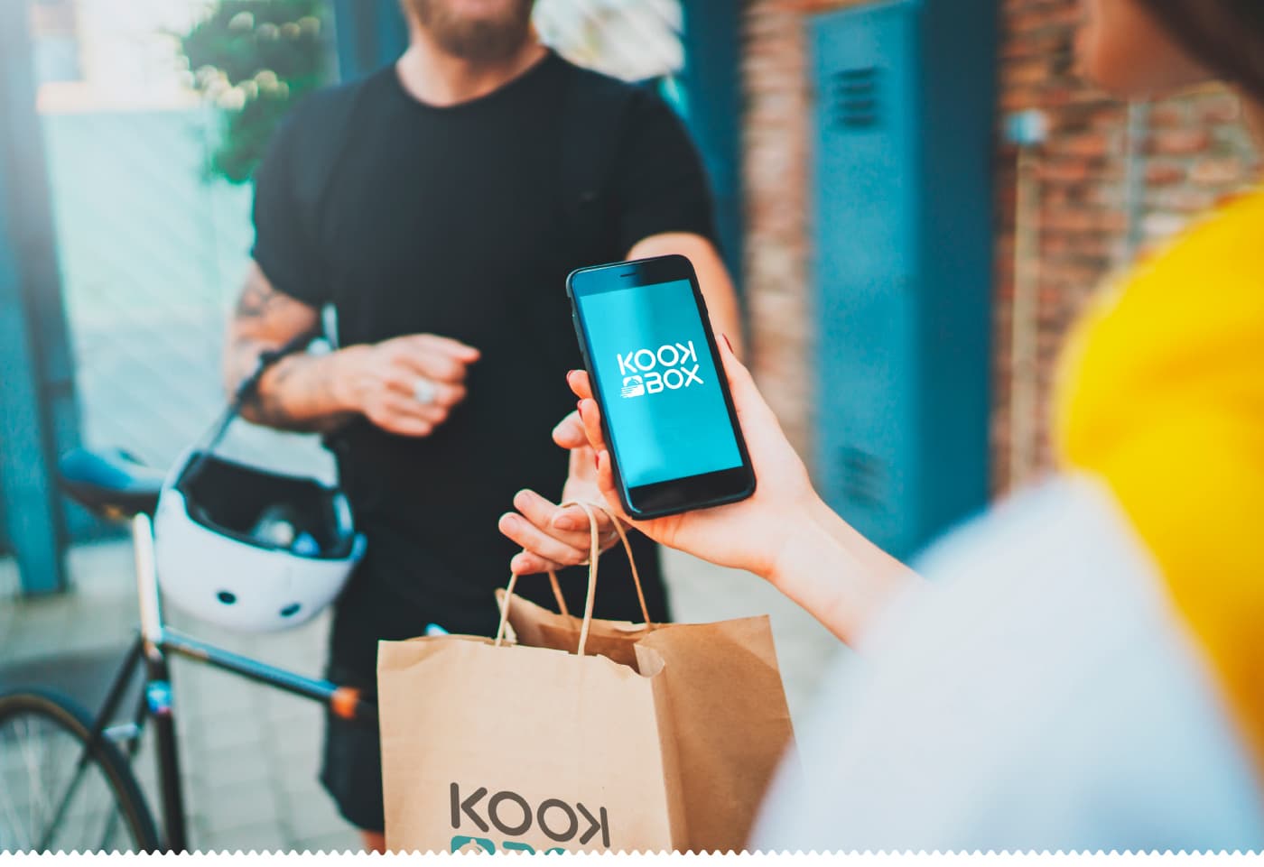 Repartidor en bicicleta entregando una bolsa de KookBox mientras la clienta muestra la app en el móvil